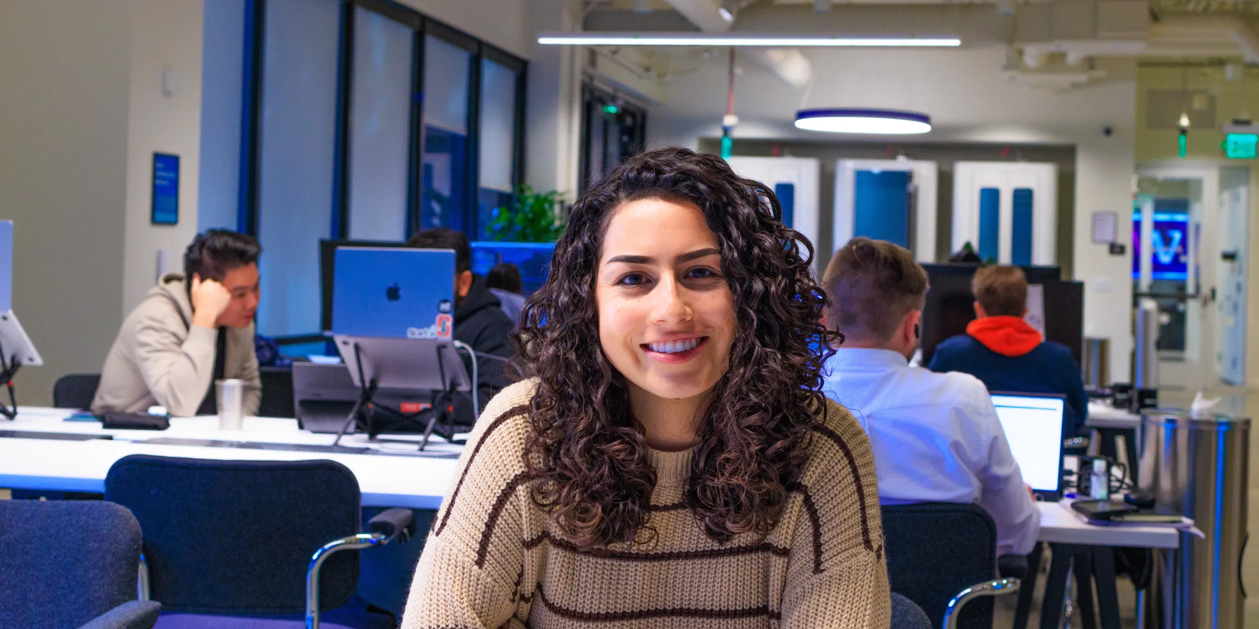 A smiling woman sitting in a modern office space with coworkers working at desks in the background, creating a collaborative and tech-driven environment. Multiple Apple computers and a bright, open workspace are visible.